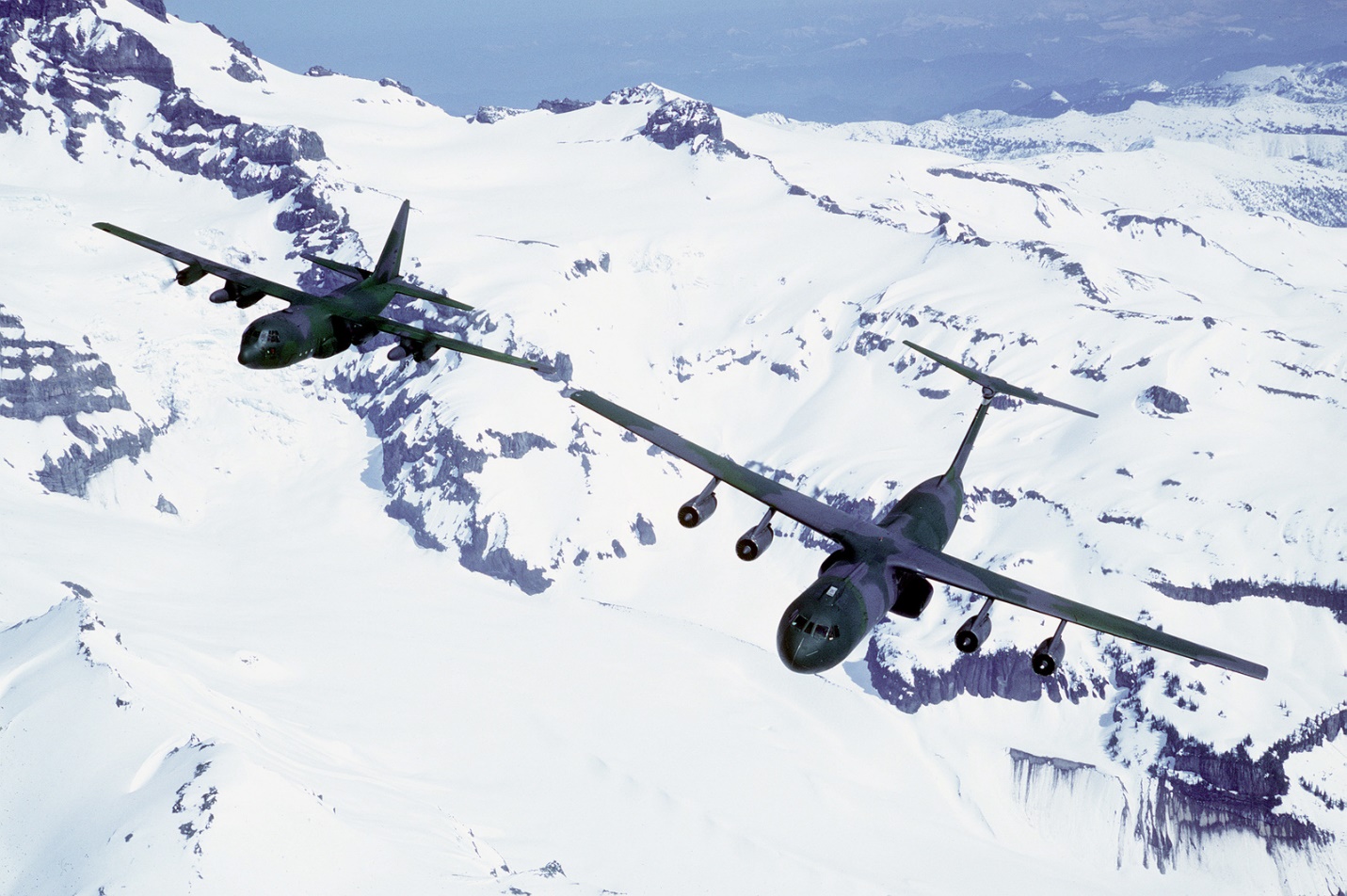An air-to-air front view of a C-141 Starlifter aircraft (right) from the 62nd Military Airlift Squadron, and a C-130 Hercules aircraft above snow covered mountains - U.S. National Archives &amp; DVIDS Public